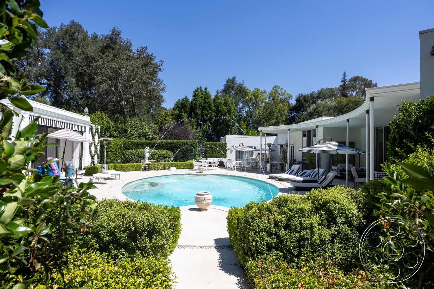Retro 295 - exterior, white stucco walls, sliding glass doors, modern outdoor furniture, concrete pool deck, oval swimming pool, surrounding trees and greenery, landscaped garden with hedges, decorative stone fountain, patio seating area with striped canopy