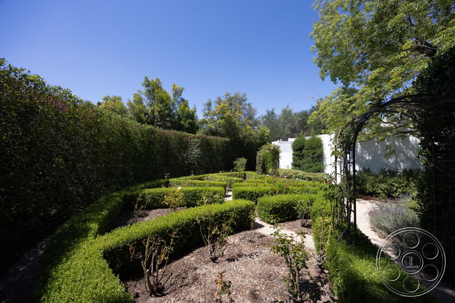 Retro 295 - exterior, white stucco walls, surrounding trees, blue sky, manicured hedges, boxwood hedges, landscape garden, gravel garden path, sunlit garden area, curved garden pathways, flowering rose bushes, metal arch trellis