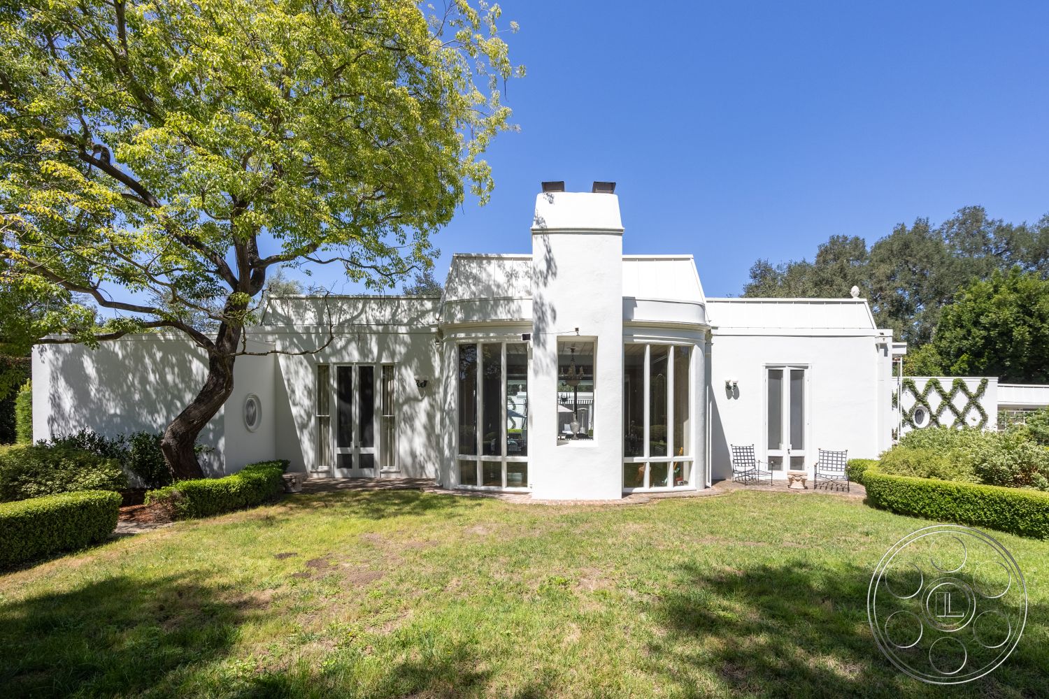 Retro 295 - exterior, white stucco walls, outdoor seating area, floor-to-ceiling windows, modern architectural style, manicured hedges, flat roof design, landscaped garden area, sunny backyard setting, curved wall section, decorative wall trellises, isolated residential location