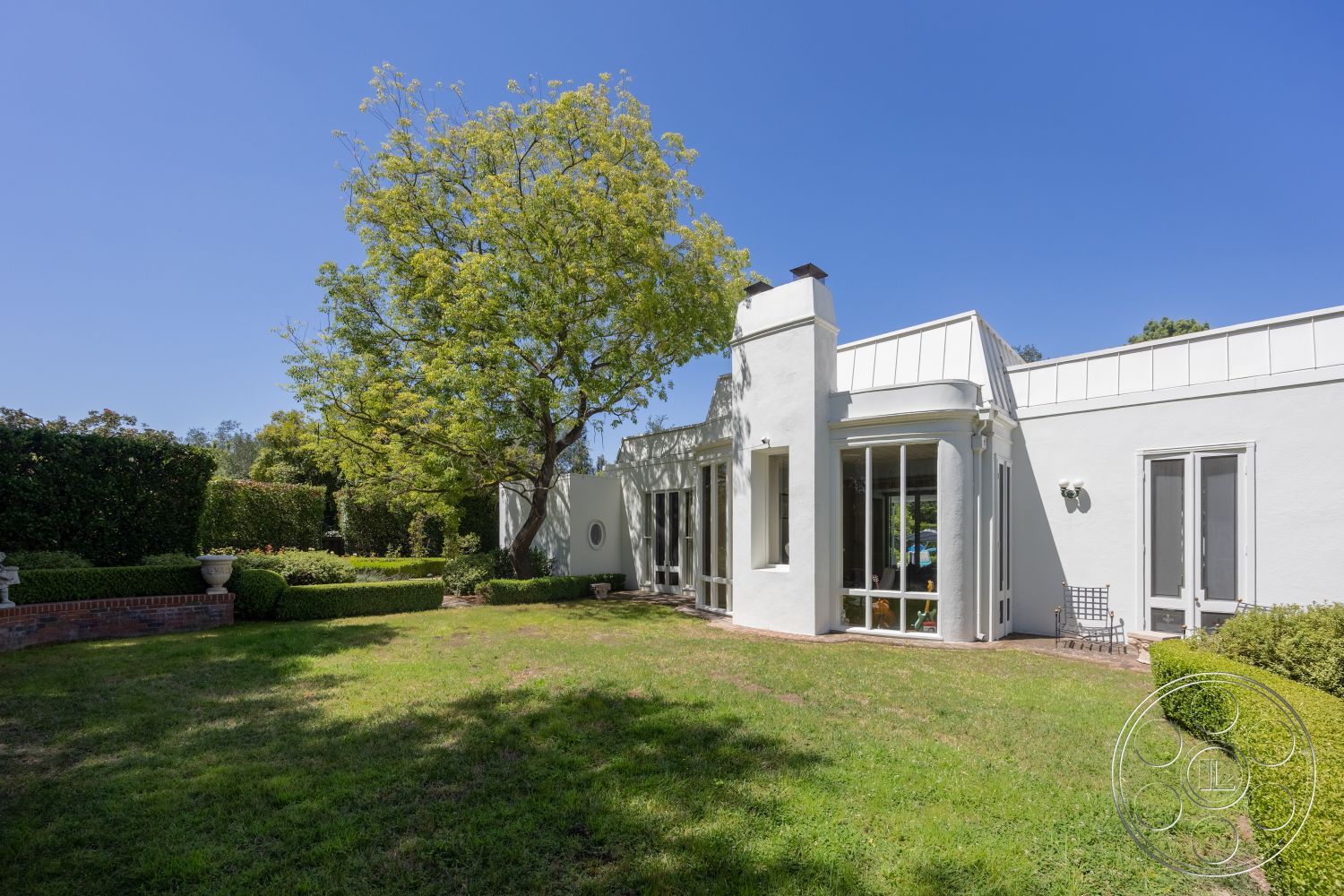 Retro 295 - exterior, white stucco walls, flat roof, large glass windows, lawn area, circular seating area, brick patio border, landscaped garden with hedges, mature tree in yard, charming outdoor ambiance