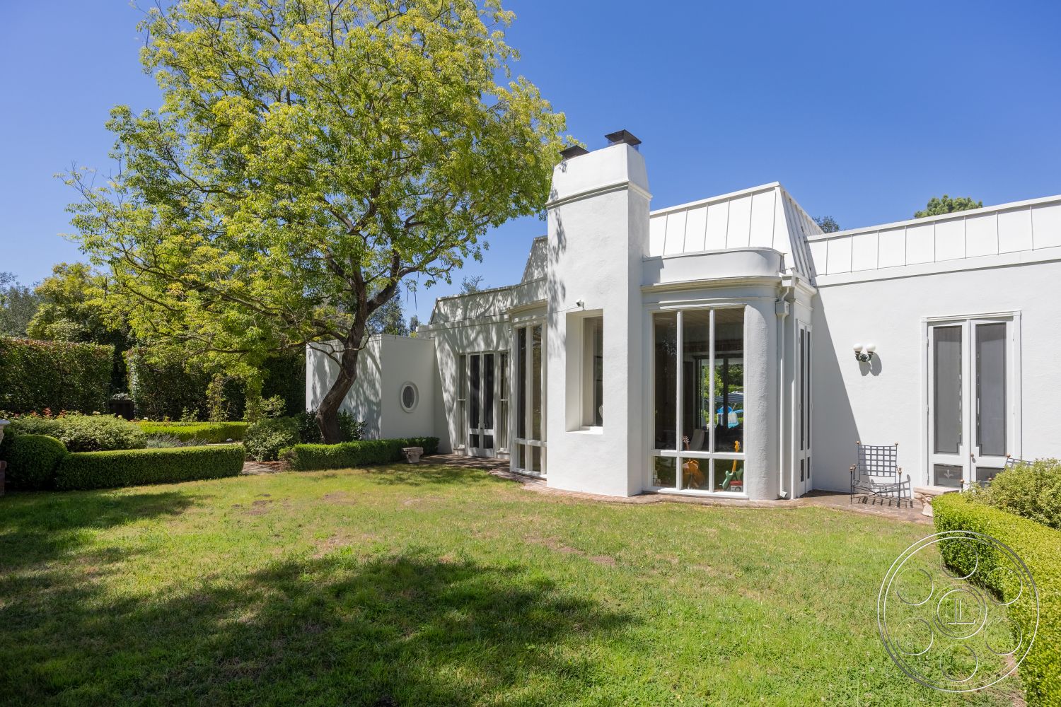 Retro 295 - exterior, landscaped garden, white stucco walls, outdoor seating area, brick patio, large trees, modern architectural style, lawn area, rectangular windows, flat roof design, sloped roof detail, hedged greenery