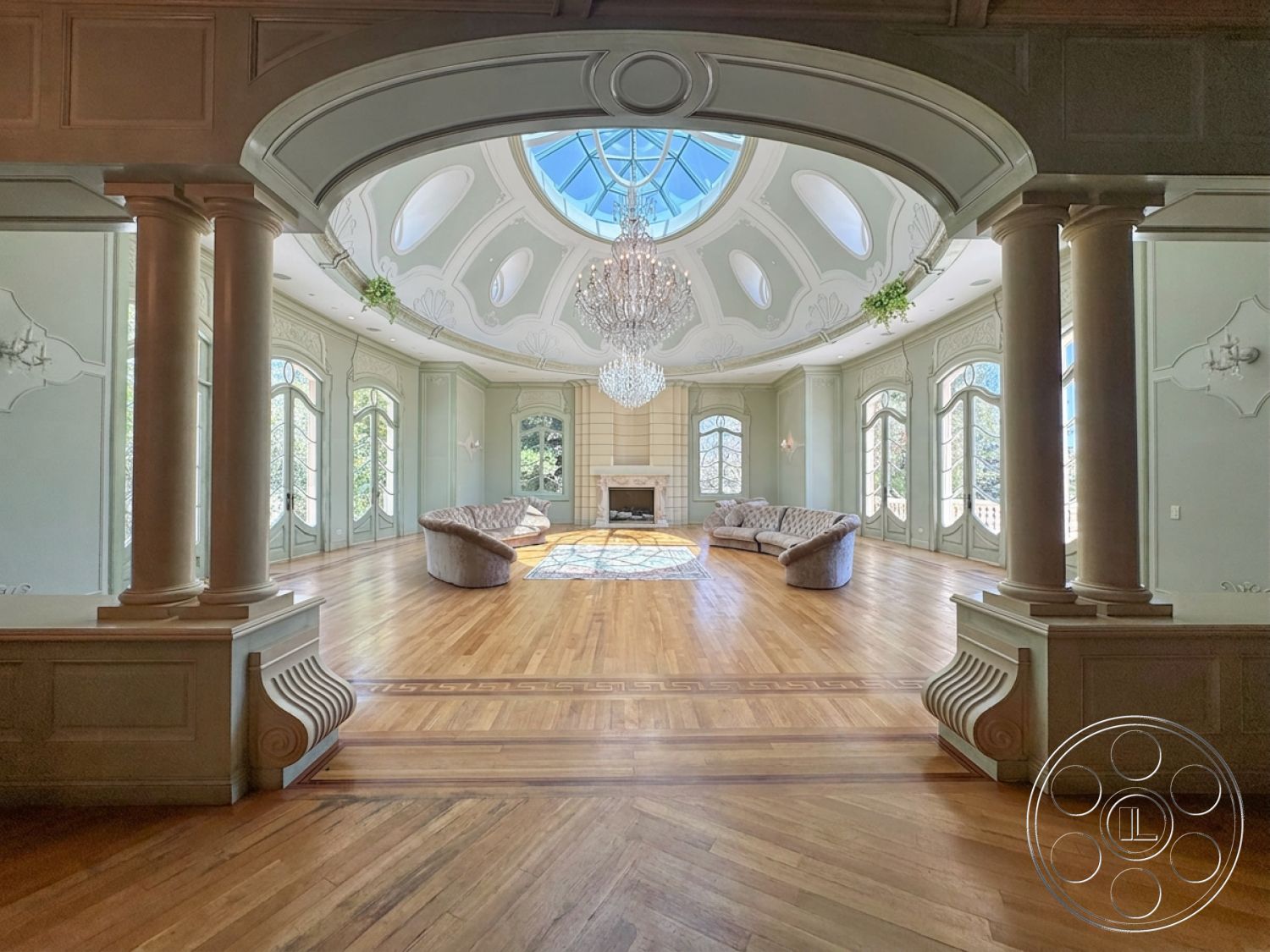 Tiffany Estate - hardwood flooring, greenery accents, area rug, exposed wooden beams, vaulted ceiling, luxurious design elements, arched windows, chandelier light fixture, decorative wall paneling, natural light from skylight, ornate ceiling moldings, elegant living room, fireplace feature wall, symmetrical room layout, classical column details, detailed wood trim, rounded sofa