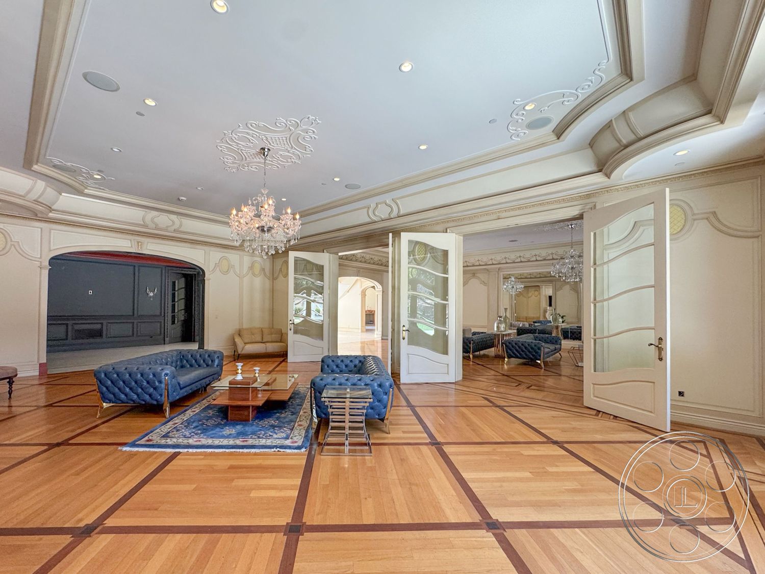 Tiffany Estate - hardwood flooring, decorative area rug, open concept living space, traditional architectural style, elegant coffee table, recessed ceiling lights, neutral painted walls, chandelier lighting fixture, double doors with glass panels, geometric patterned hardwood floor, plaster ceiling with intricate molding, upholstered blue leather sofas, ornamental wall details