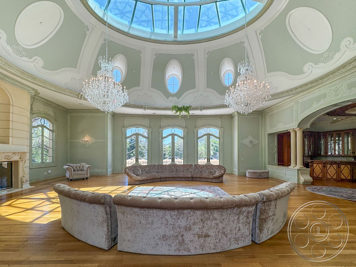 Tiffany Estate - hardwood flooring, large arched windows, textured wall treatments, natural light from skylight, open living space layout, decorative ceiling medallion, chandelier lighting fixture, intricate wall moldings, rounded velvet furniture, circular dome ceiling, plaster ceiling details, granite fireplace surround, broad circular area rug, built-in cabinetry in background
