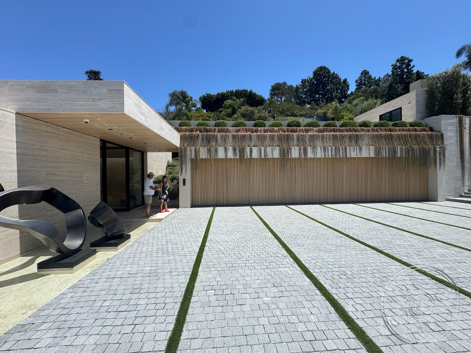 Chalon Estate - exterior, modern architecture, floor-to-ceiling windows, landscaped front yard, wooden garage door, textured stone walls, cobblestone driveway, green grass accents, flat roof design, lush greenery landscaping, rectangular driveway layout, exposed aggregate patio, metal sculpture artwork