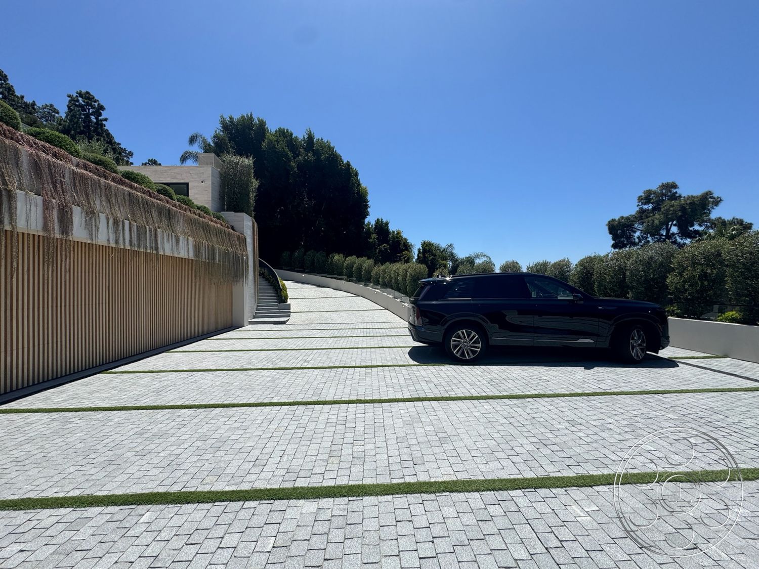 Chalon Estate - exterior, paved driveway, modern architecture, lush greenery, curved driveway, sloped driveway, clear blue sky, landscaped hedges, wooden retaining wall, gray stone pavers, lawn inlay strips