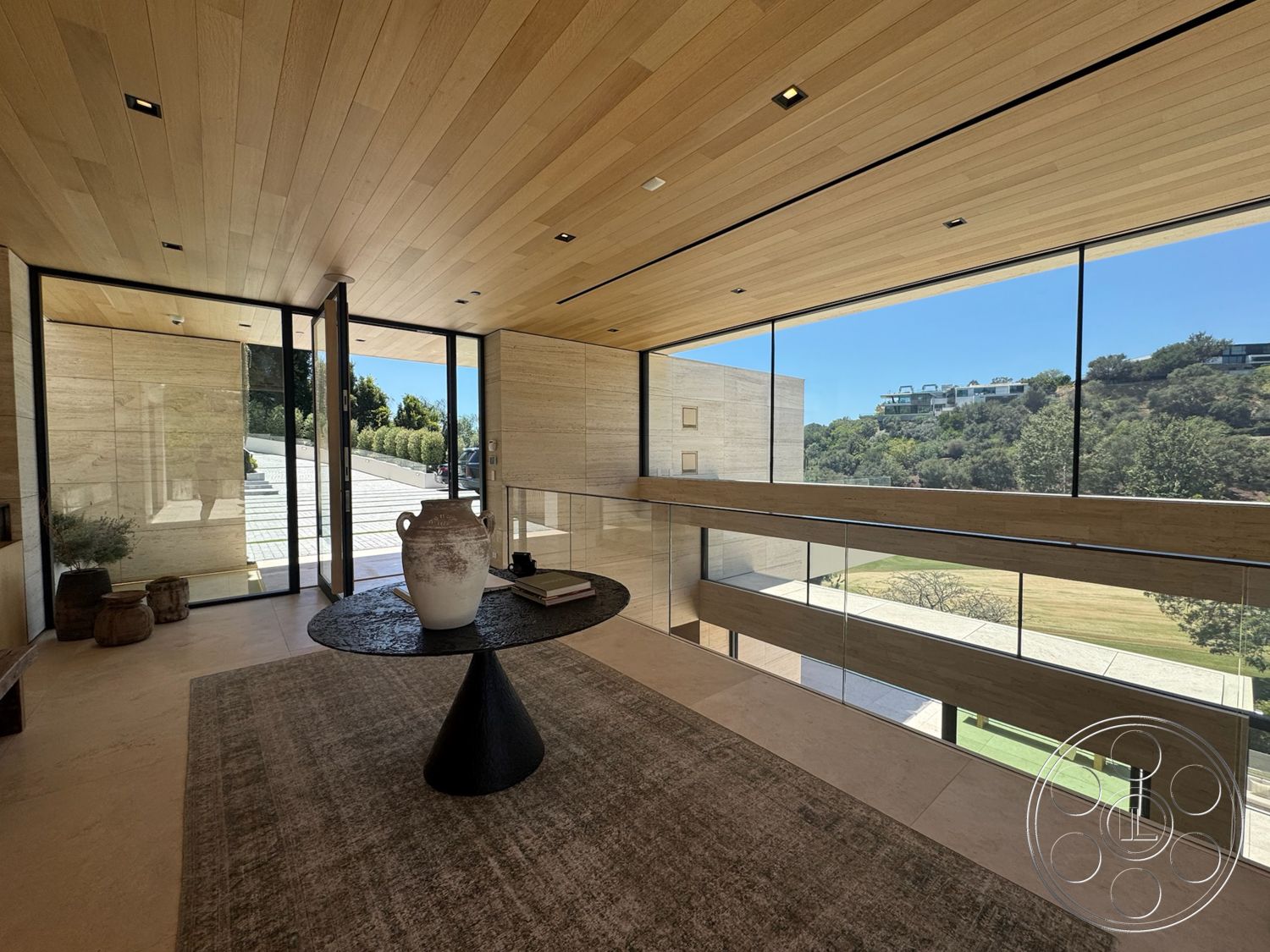 Chalon Estate - neutral color palette, natural light, stone accent wall, textured area rug, concrete flooring, spacious living area, contemporary architecture, minimalist decor style, floor-to-ceiling glass windows, suburban hillside setting, open entryway design, wood ceiling paneling, modern circular coffee table, lush hillside view, elegant vase decor