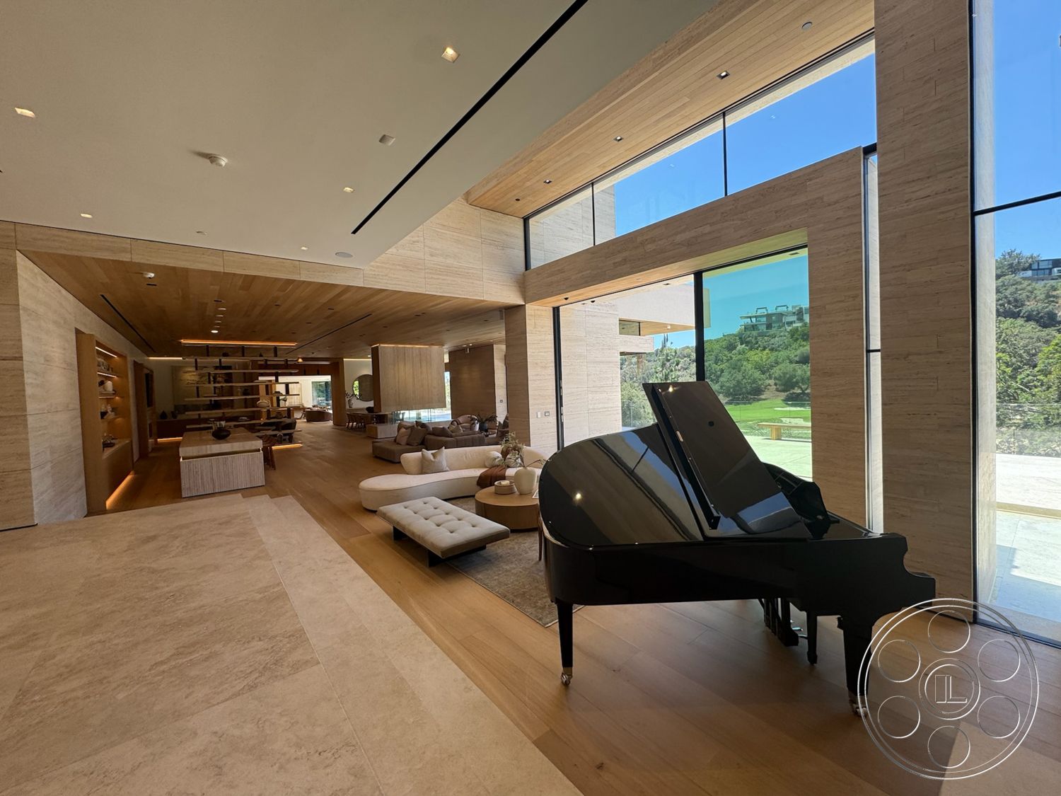 Chalon Estate - minimalist decor, stone accent wall, open floor plan, modern furniture, black grand piano, light wood paneling, neutral color scheme, designer lighting fixtures, large windows with garden view, tile living room flooring, natural wood ceiling beams, glass wall openings, sectional sofa with throw pillows, custom wood shelving, coffee table with decorative accessories