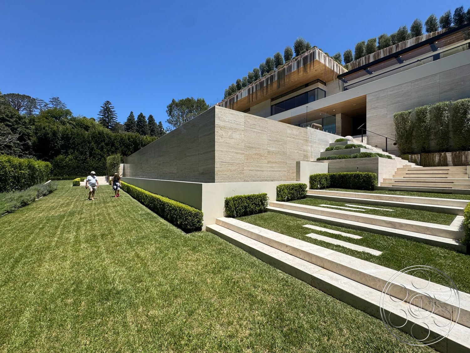 Chalon Estate - exterior, modern architectural style, concrete retaining wall, sunny hillside location, natural stone wall cladding, flat grassy area, horizontal wood roof overhang, landscaped garden with trimmed hedges, formal lawn area, rectangular stone steps, elevated outdoor space, structured planting beds