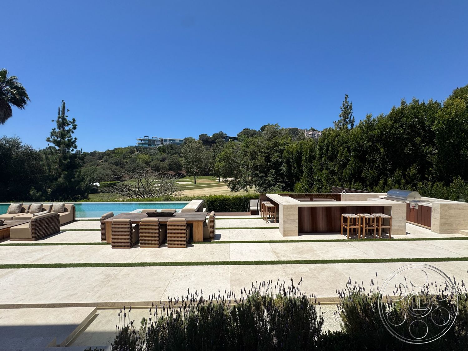 Chalon Estate - exterior, rectangular swimming pool, stainless steel grill, clear blue sky, wooden outdoor furniture, tall pine trees, lush green landscaping, stone patio flooring, built-in outdoor kitchen, bar height wooden counter
