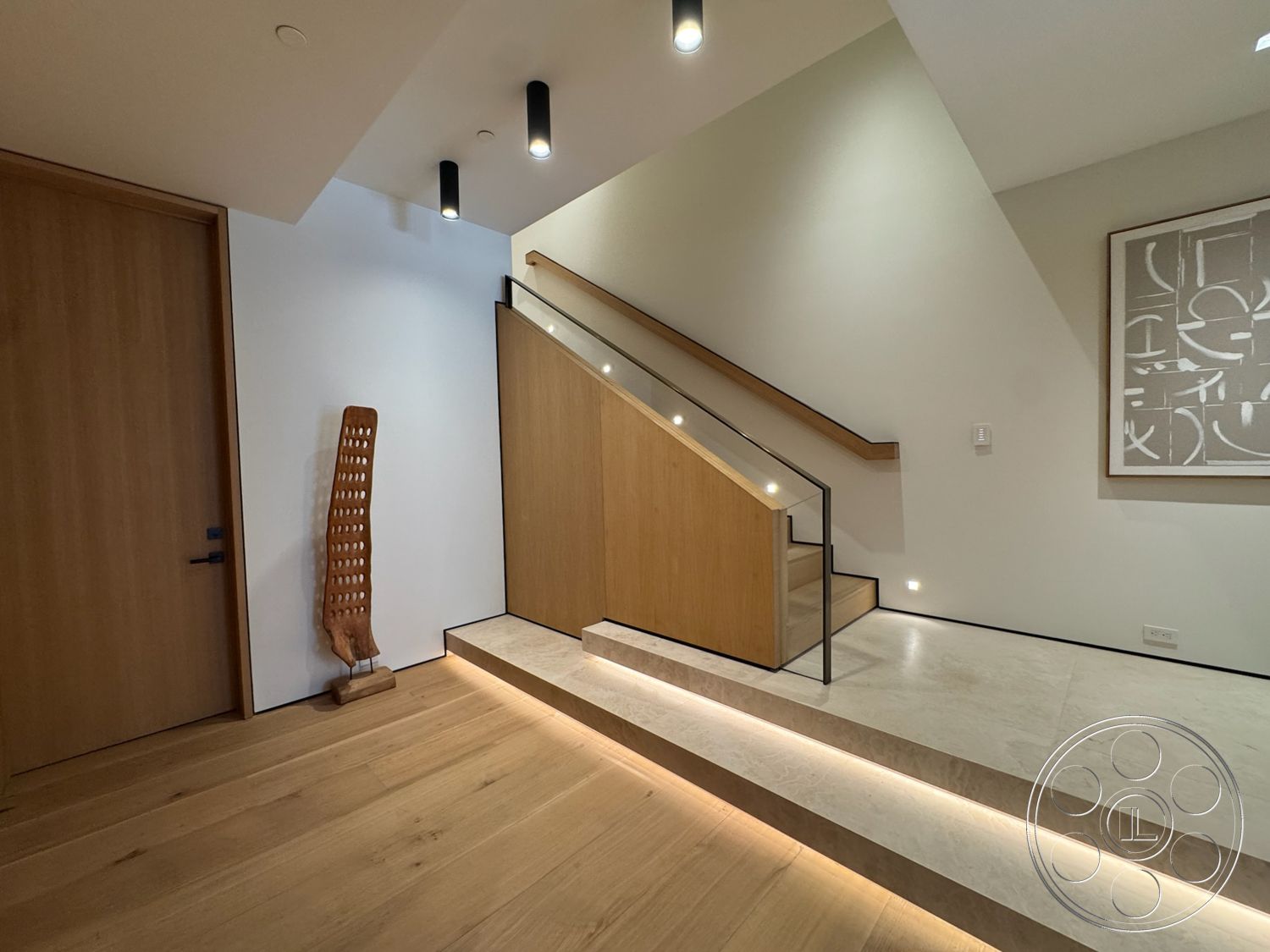 Chalon Estate - light wood flooring, textured accent wall, modern art wall decor, neutral painted walls, exposed wood handrail, marble entryway flooring, dark wood front door, interior wooden staircase, glass stair railings, high ceiling lighting fixtures