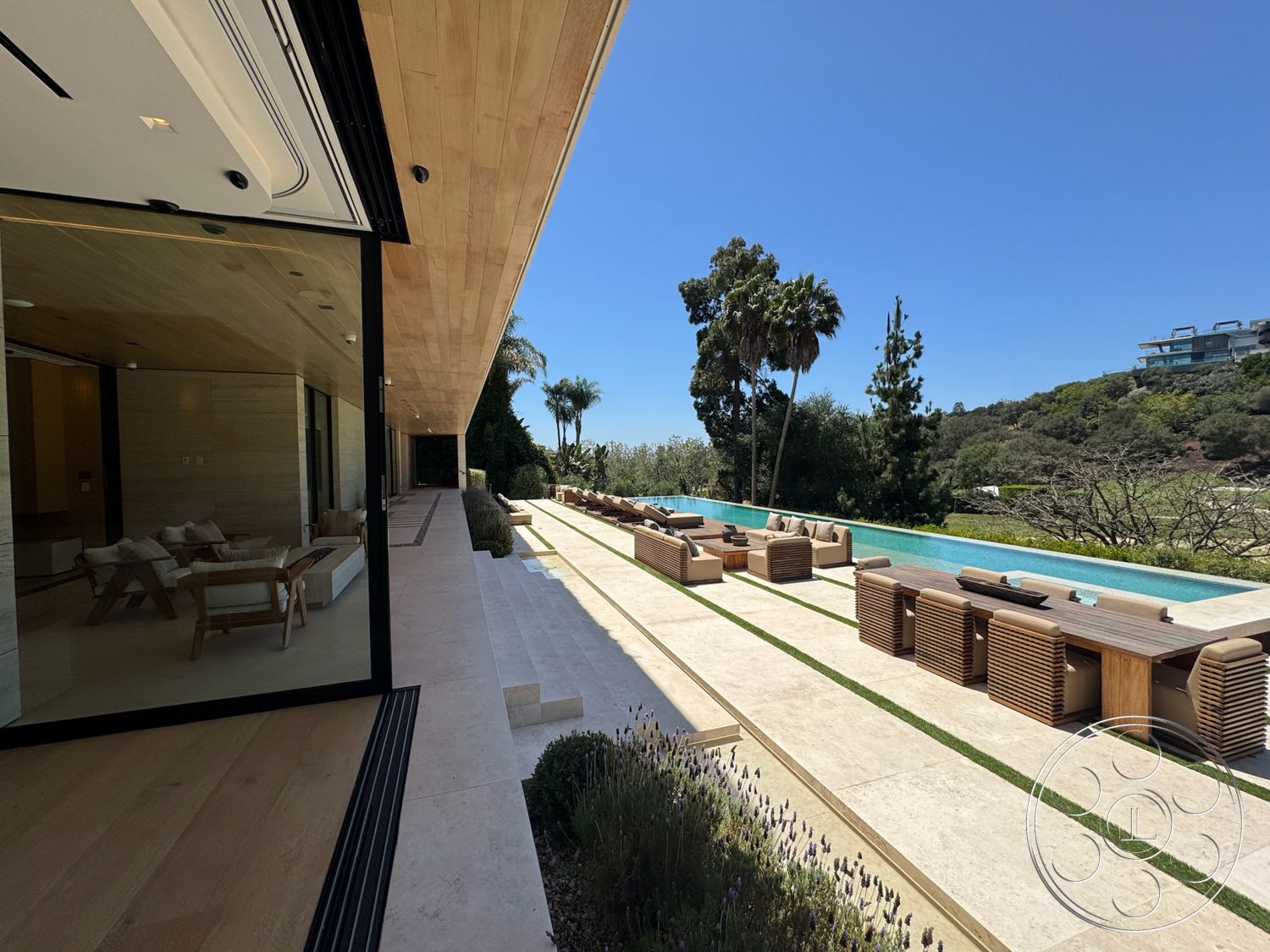 Chalon Estate - exterior, glass sliding doors, outdoor lounge area, clear blue sky, natural stone path, palm trees in landscape, rectangular infinity pool, surrounding hillside view, smooth concrete patio flooring, desert landscaping with drought-resistant plants, wood beam soffit, level grass strips between pool and seating area, high-end modern furniture with slatted detailing