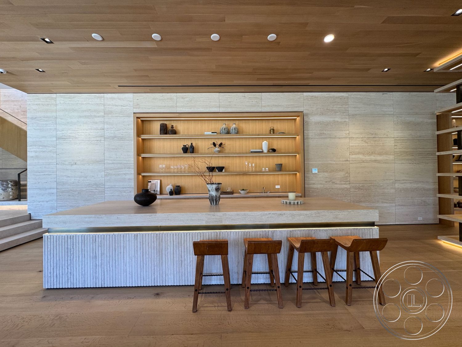 Chalon Estate - natural wood bar stools, modern minimalist design, textured stone wall panels, spacious open floor plan, plank wood flooring, light oak wood ceiling, white marble accent wall, light beige marble kitchen island, built-in wooden shelf with LED lighting, stainless steel kitchen fixtures