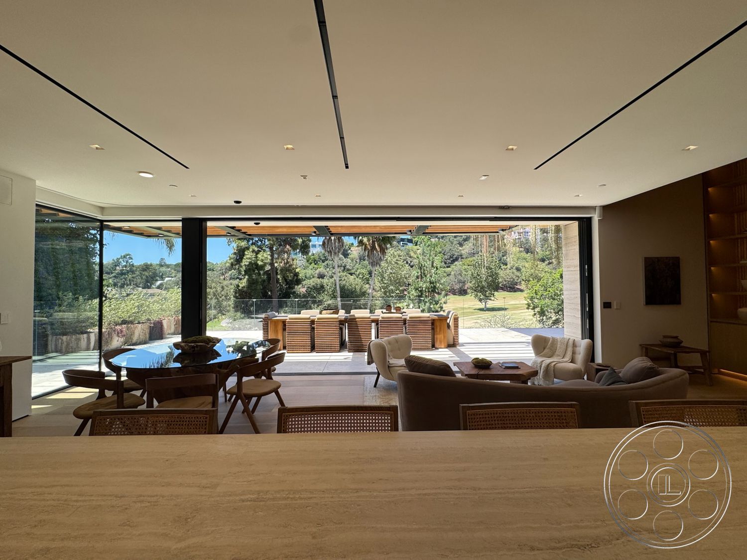 Chalon Estate - neutral color palette, open floor plan, glass sliding doors, modern coffee table, light wood cabinetry, floor-to-ceiling windows, round dining table, textured wall treatment, wooden outdoor furniture, recessed ceiling lights, wood dining chairs, natural light-filled space, lush greenery setting, contemporary dining room, woven accent chairs, expansive nature view, stone kitchen countertop, minimalist living room, outdoor deck area, secure glass railing