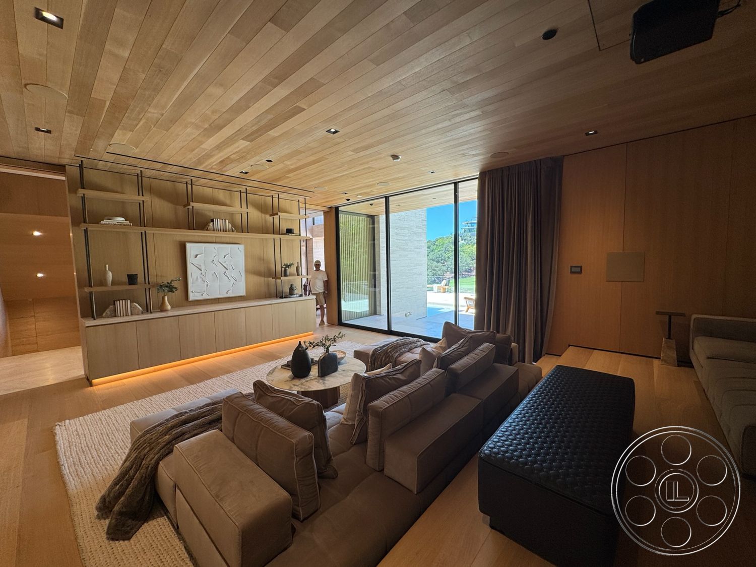 Chalon Estate - hardwood flooring, minimalist design elements, large floor-to-ceiling windows, textured beige area rug, open living space layout, contemporary furniture style, built-in wooden shelves, natural light abundance, light wood ceiling panels, plush taupe sectional sofa, dark leather ottoman, neutral tone wall paneling, decorative white wall art
