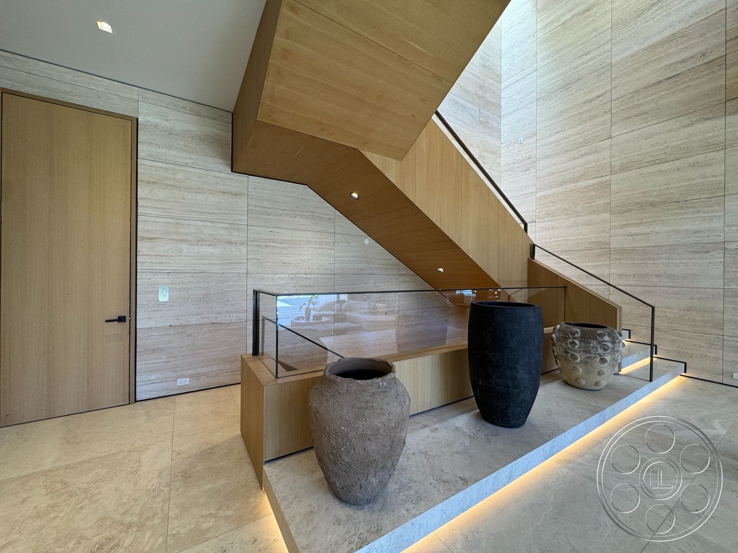 Chalon Estate - recessed lighting, minimalist design, modern entryway, glass railing, wooden staircase, open space concept, textured stone walls, decorative ceramic vases, light hardwood treads, tan stone flooring, large wooden panel door