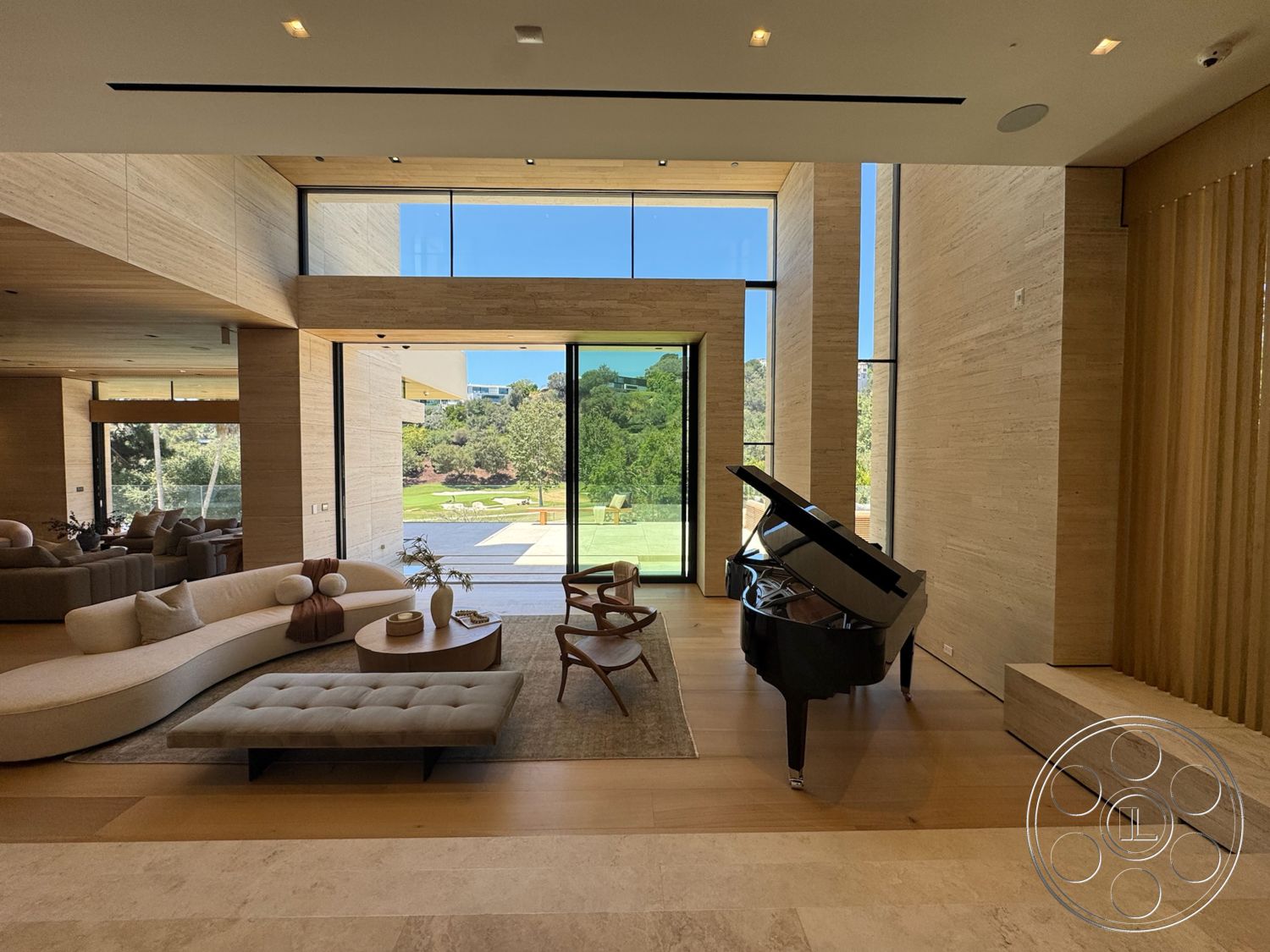 Chalon Estate - minimalist design, floor-to-ceiling windows, black grand piano, modern architectural style, curved sectional sofa, concrete patio flooring, wood beam ceiling, neutral-toned area rug, natural light filled space, landscape view through windows, wood panel walls, tufted coffee table, light-colored hardwood flooring