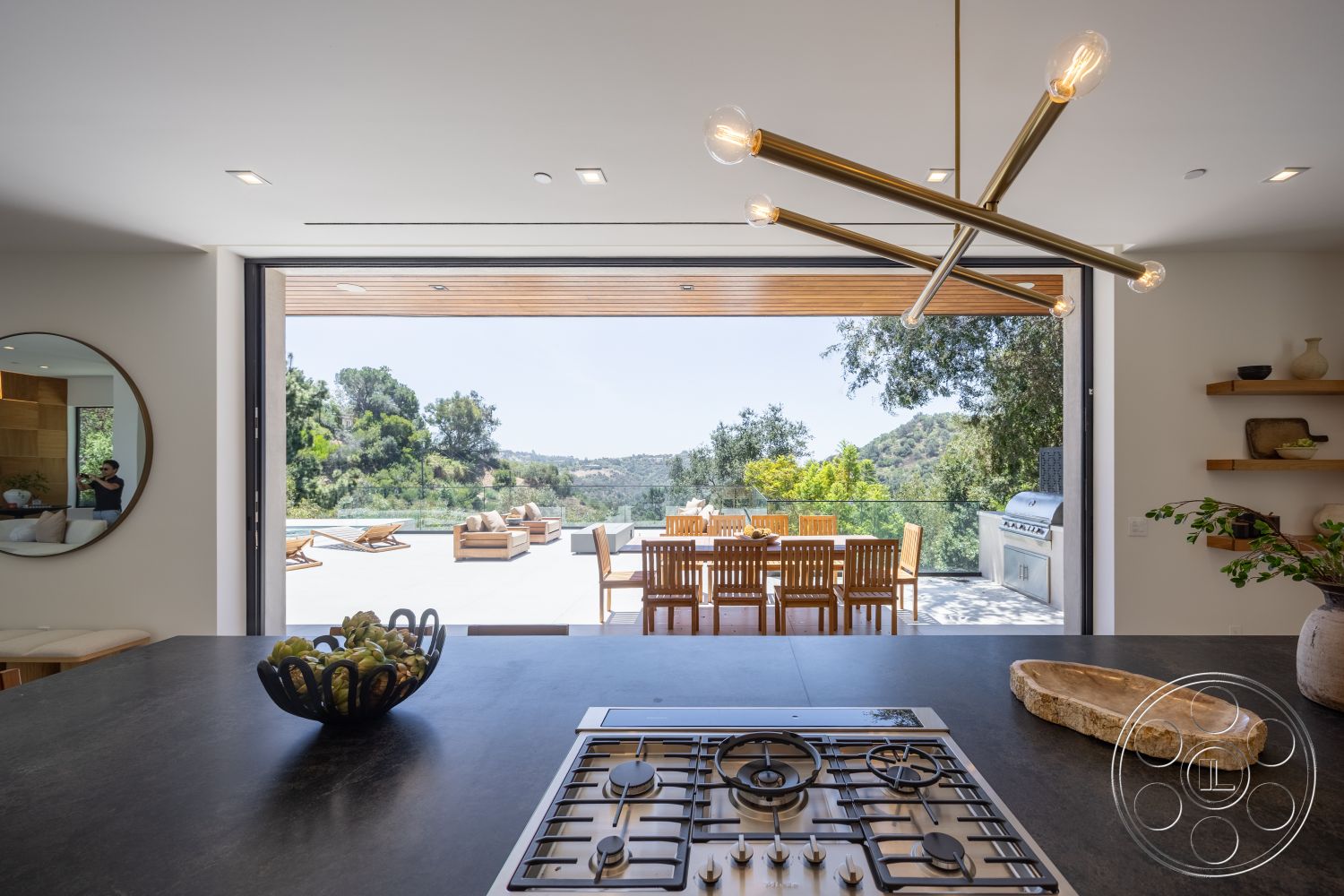 Canyon House - open concept layout, wooden ceiling beams, natural wood dining table, wooden shelving units, white painted walls, floor-to-ceiling glass sliding doors, outdoor patio area with lounge chairs, stainless steel gas cooktop, interior kitchen with concrete countertops, mountain view from kitchen, elegant pendant lighting fixtures, minimalist decorative bowl