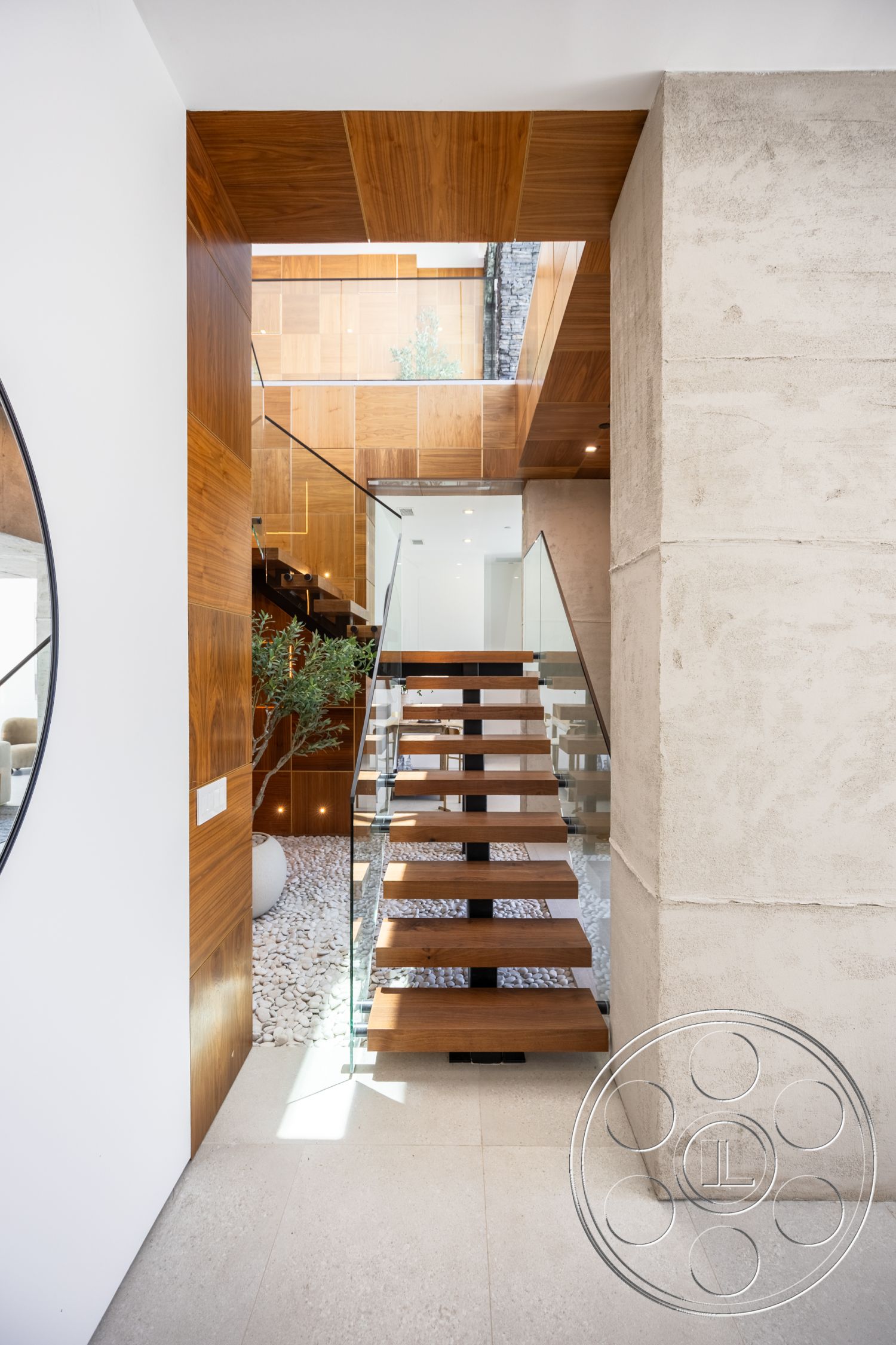 Canyon House - wood ceiling, concrete walls, modern architectural design, natural light entry, floating wooden staircase, glass stair railings, polished stone floor tiles, pebble garden floor, exposed wooden paneling, circular mirror wall feature, indoor plant area