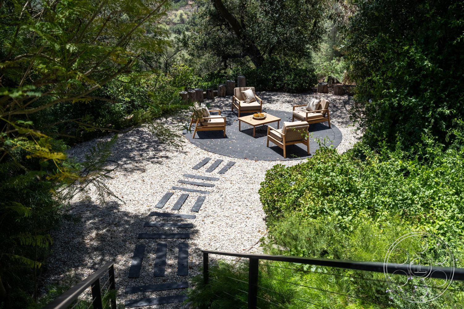Canyon House - outdoor seating area, wooden patio furniture, circular seating area, gravel garden flooring, rustic outdoor table, natural stone stepping stones, lush green landscape setting, slate patio surface, surrounded by dense foliage, exterior garden environment