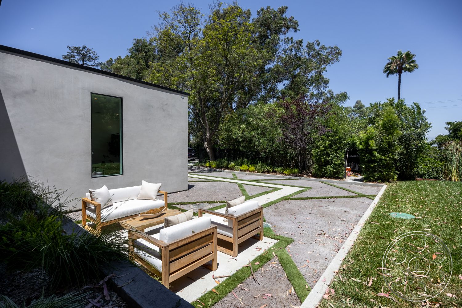 Canyon House - exterior, landscaped garden, palm trees, tropical plants, concrete driveway, tall trees, stucco walls, suburban backyard, natural grass lawn, large glass window, paved patio area, wood outdoor seating area, rectangular outdoor chairs, geometric patterns in concrete