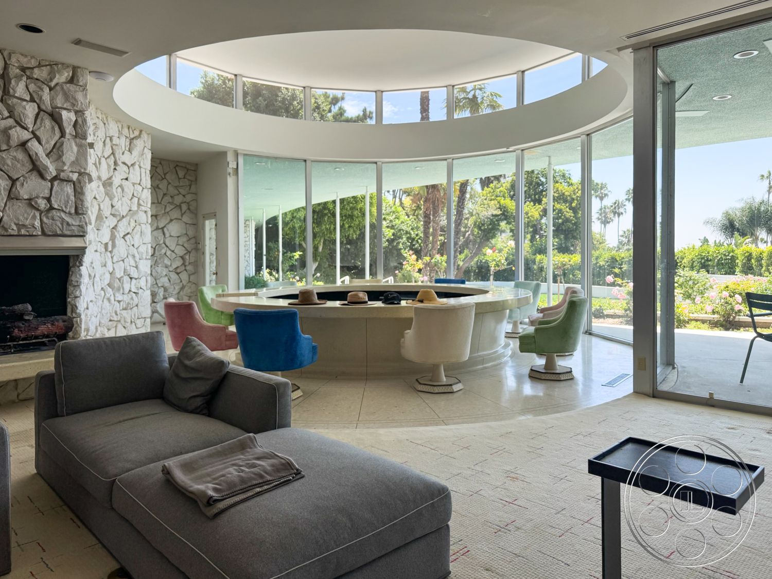 MCM 28 - exterior, open floor plan, concrete floor, landscaped garden view, round dining table, natural light through large windows, modernist architectural style, rounded glass walls, exposed stone fireplace wall, textured white stone accents, colorful upholstered chairs, sitting area with gray sectional sofa