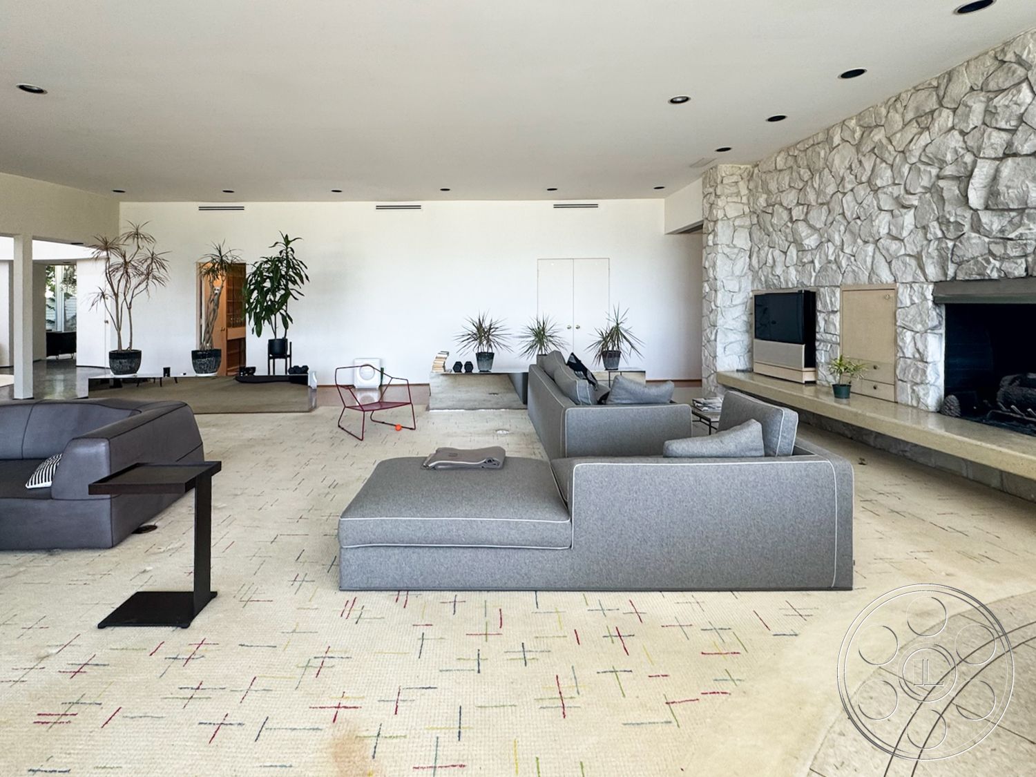 MCM 28 - neutral color palette, minimalist coffee table, mid-century modern style, stone feature wall, open floor plan layout, modern sectional sofa, decorative indoor plants, exposed ceiling lights, large windows with natural light, concrete fireplace surround, carpet living room flooring