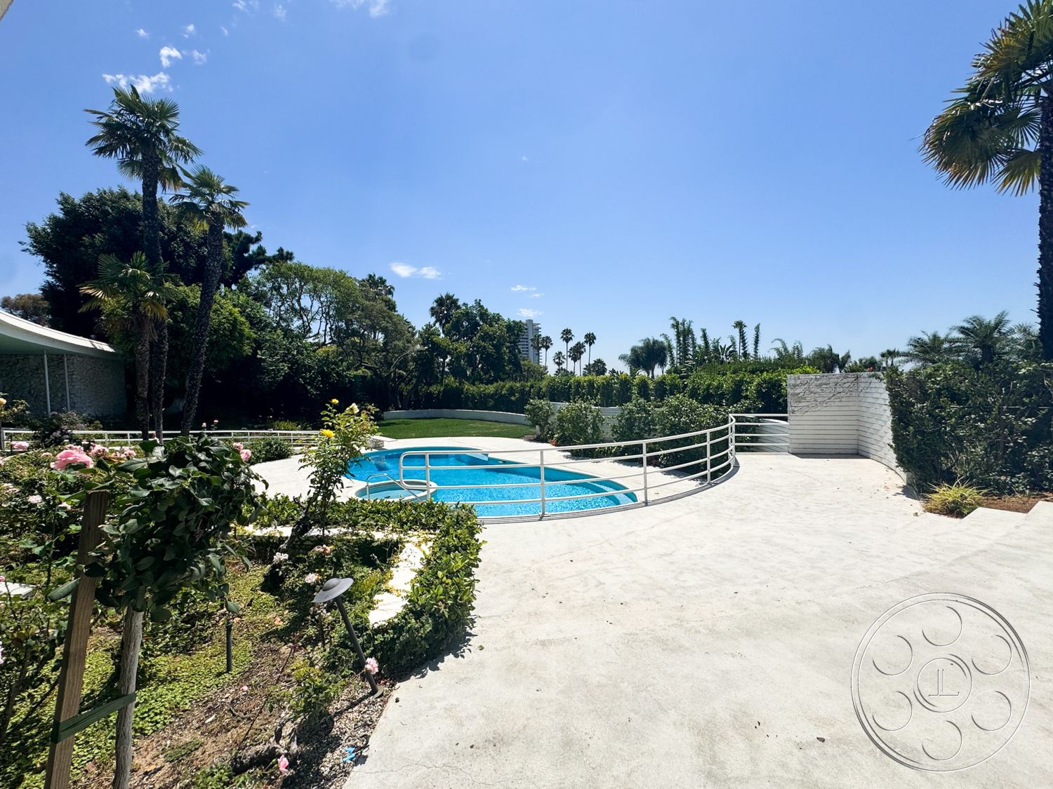 MCM 28 - exterior, lush greenery, palm trees, rectangular swimming pool, clear blue sky, white brick wall, blue pool water, sunny outdoor setting, concrete patio flooring, landscaped garden with flowers, plantings of shrubs and hedges, curved poolside railing