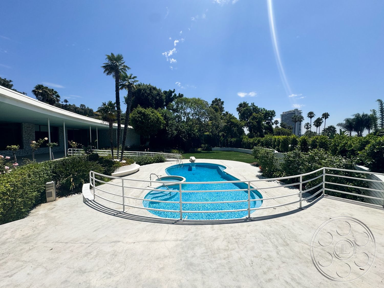 MCM 28 - exterior, landscaped garden, lush green lawn, tropical palm trees, concrete pool deck, mid-century modern architecture, curved pool, sunny outdoor setting, modern home design, concrete patio surface, single-story residence, glass railing fence, blue mosaic pool tile