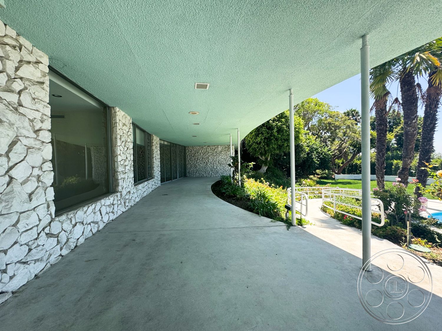 MCM 28 - exterior, floor-to-ceiling windows, open outdoor space, textured plaster ceiling, curved walkway, concrete patio flooring, stone exterior walls, surrounding garden landscape, palm trees in the environment