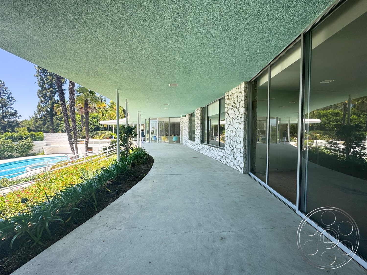 MCM 28 - exterior, stone accent wall, floor-to-ceiling windows, glass facade, modern architectural style, concrete walkway, swimming pool view, outdoor living area, palm trees in background, landscaped garden beds, tropical garden setting, curved roof overhang, exposed aggregate finish