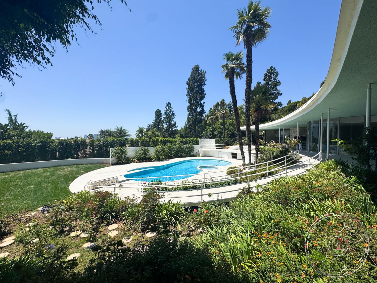 MCM 28 - exterior, white stucco walls, lush greenery, palm trees, floor-to-ceiling windows, in-ground swimming pool, rectangular pool shape, open-air layout, curved roof design, landscaped garden area, stone garden path, large outdoor terrace, concrete pool surround, sloping hillside property