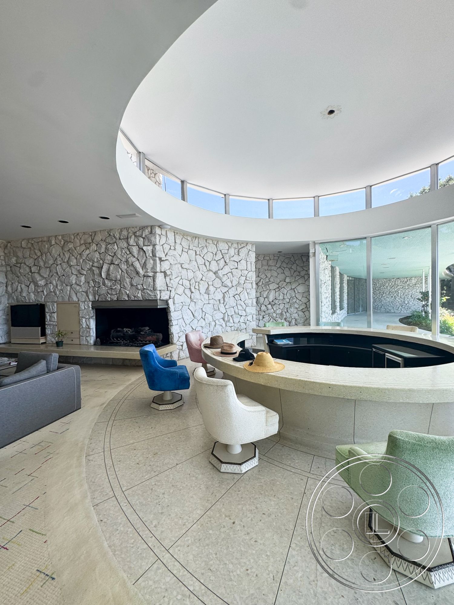 MCM 28 - large windows, natural light, open floor plan, upholstered dining chairs, circular seating arrangement, contemporary design elements, curved ceiling, stone fireplace surround, modernist architectural style, exposed stone feature wall, tile living room flooring, rounded bar counter