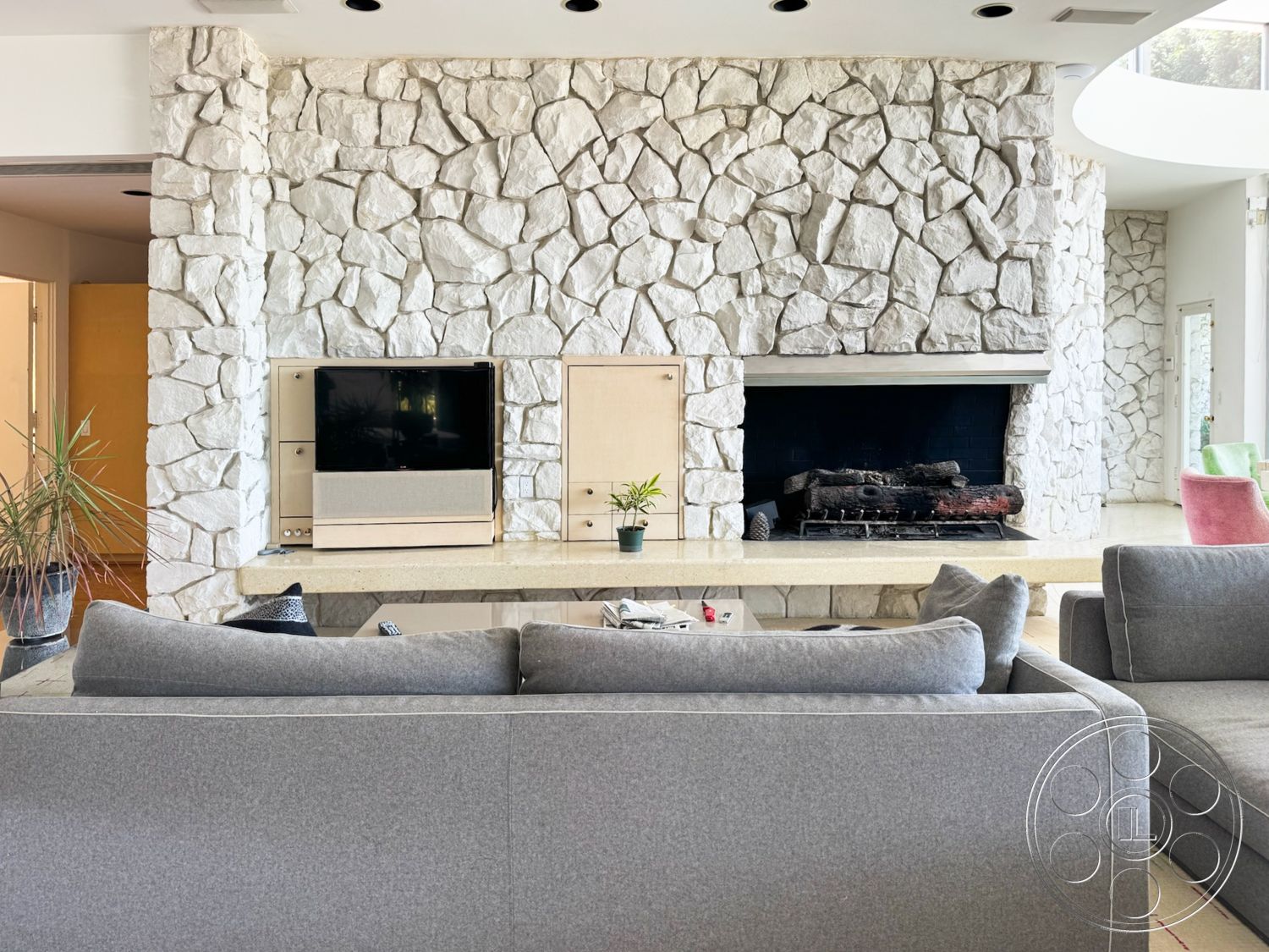 MCM 28 - minimalist decor, large windows for natural light, gray sectional sofa, stone feature wall, potted indoor plants, smooth ceiling with recessed lighting, built-in media console, white stacked stone fireplace, beige quartz coffee table, dark wood fireplace surround