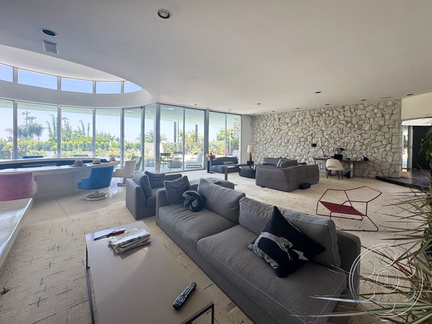 MCM 28 - neutral color palette, indoor-outdoor flow, large floor-to-ceiling windows, lush greenery visible through windows, open floor plan living area, natural light-filled spaces, curved ceiling design, exposed stone feature wall, contemporary home decor, colorful accent chairs, soft upholstered sectional sofa, concrete floor with a textured finish, modern coffee table with a minimalist design