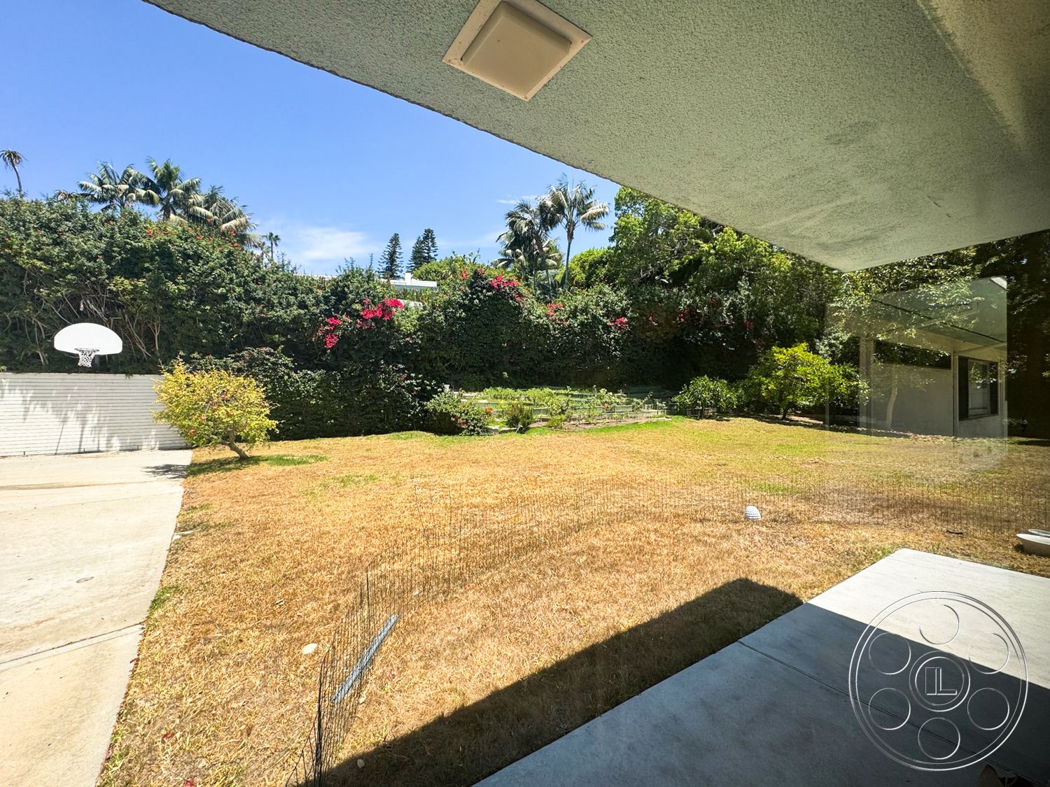 MCM 28 - exterior, fenced garden area, flowering shrubs, basketball hoop, concrete driveway, tropical landscaping, grassy lawn area, patio overhang, partial wall enclosure, wooden garden fixtures