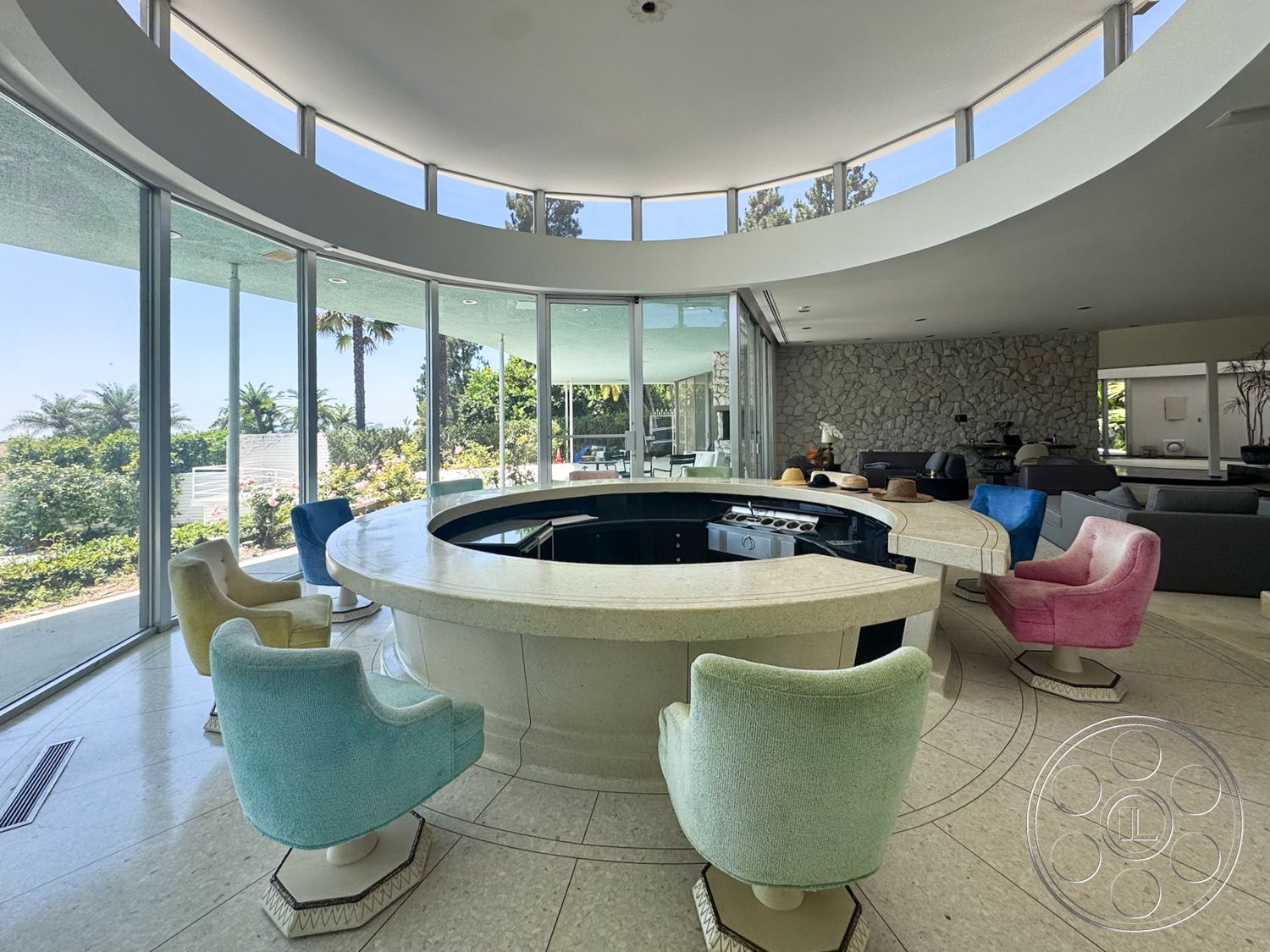MCM 28 - textured stone wall, open floor plan, modern architectural style, polished marble flooring, large floor-to-ceiling windows, lush garden view, natural light-filled space, palm tree landscaping, round layout, curved bar with stone countertop, colorful upholstered swivel chairs