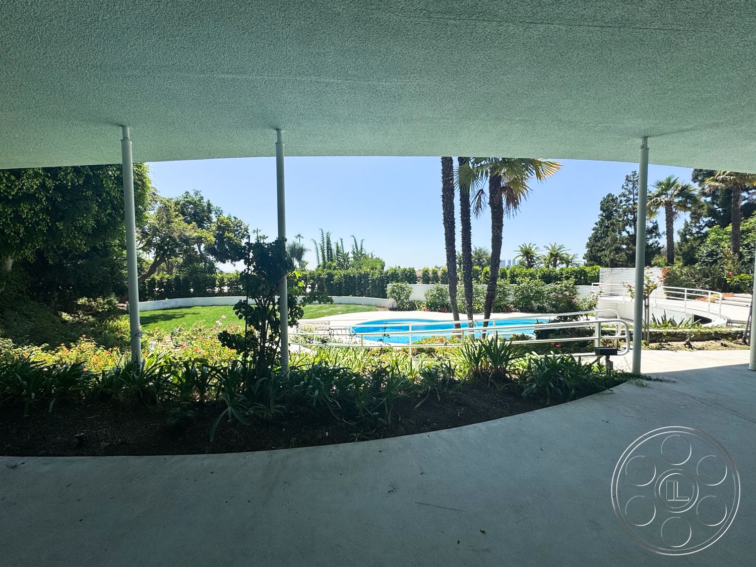 MCM 28 - exterior, palm trees, rectangular swimming pool, open outdoor space, sunny setting, curved garden beds, bush landscaping, landscaped garden area, lush green foliage, concrete patio floor, white pool railing, steps leading to pool area