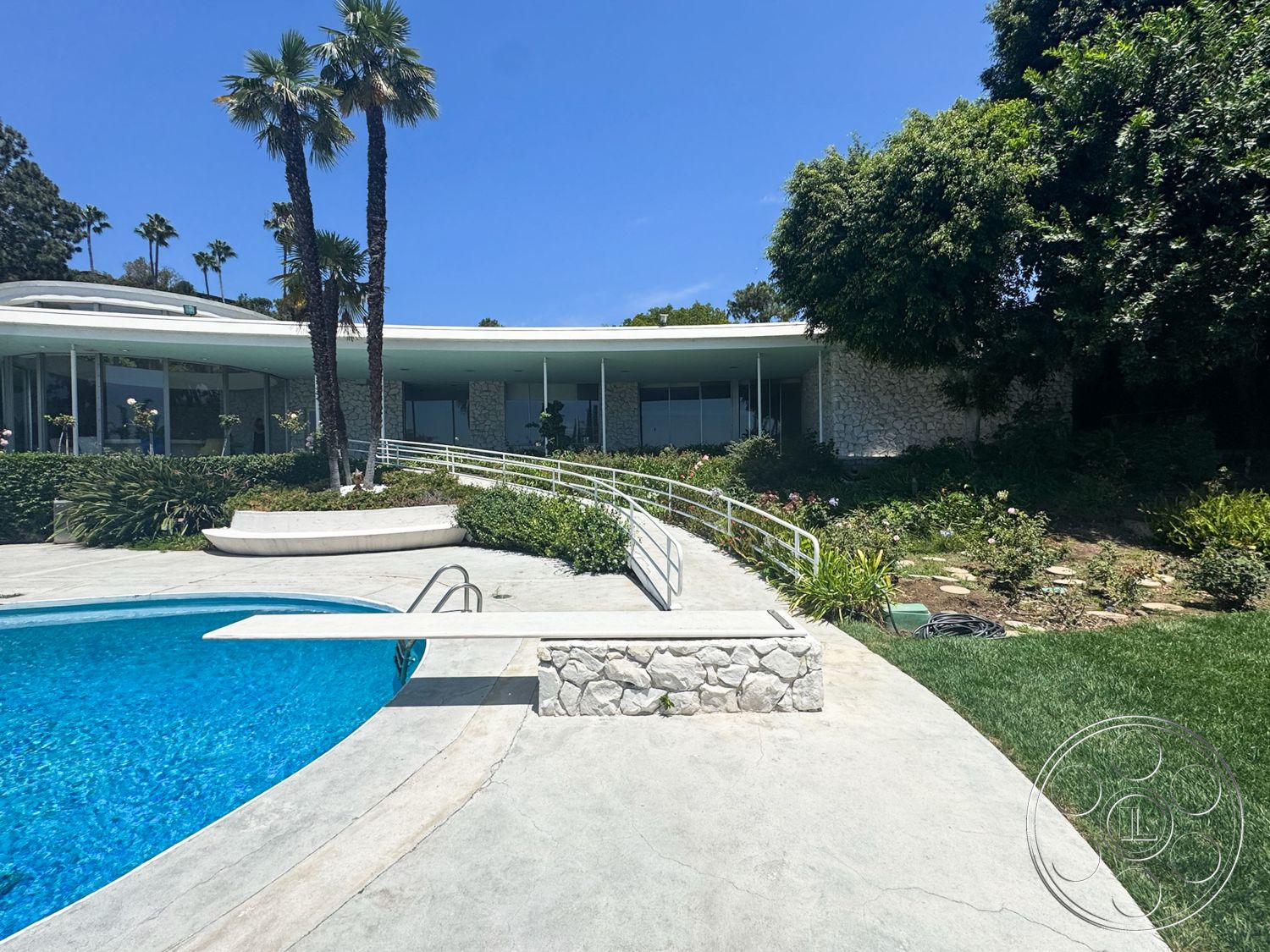MCM 28 - exterior, landscaped garden, palm trees, large glass windows, rectangular swimming pool, green lawn, curved walkway, metal handrail, exposed stone wall, mid-century modern architecture, sloping hillside, stone pool deck, concrete patio flooring, solid stone poolside edge