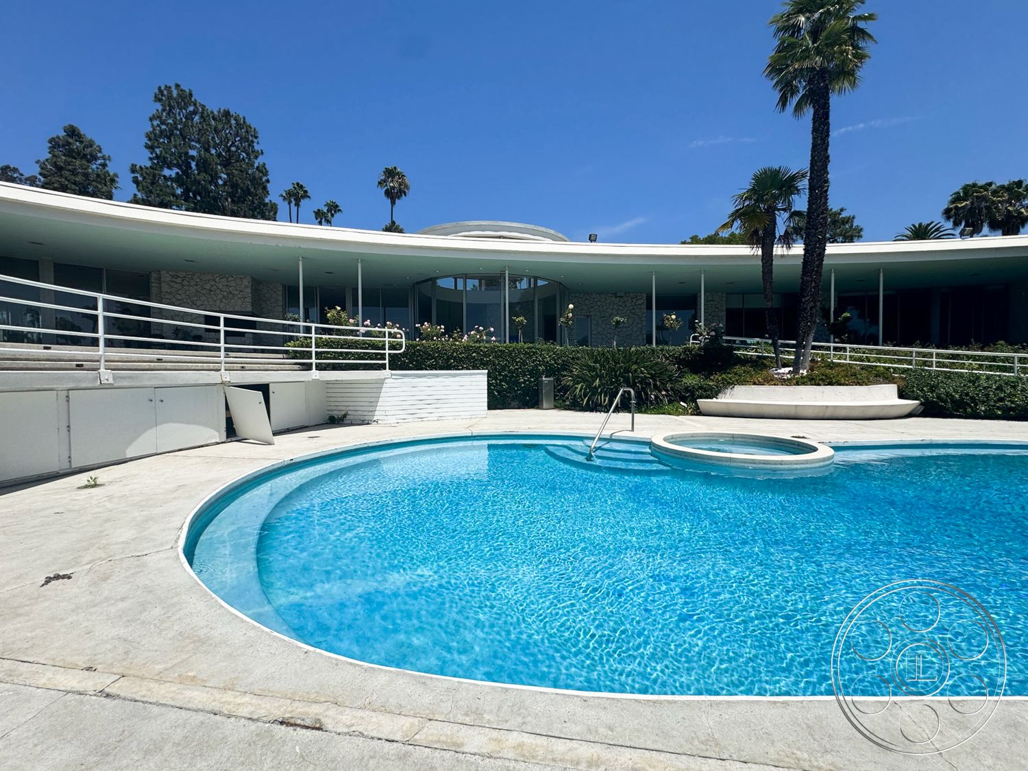 MCM 28 - exterior, palm trees, glass sliding doors, concrete pool deck, stone feature wall, mid-century modern architecture, curved swimming pool, landscaped garden area, white stucco exterior walls, patio with railing