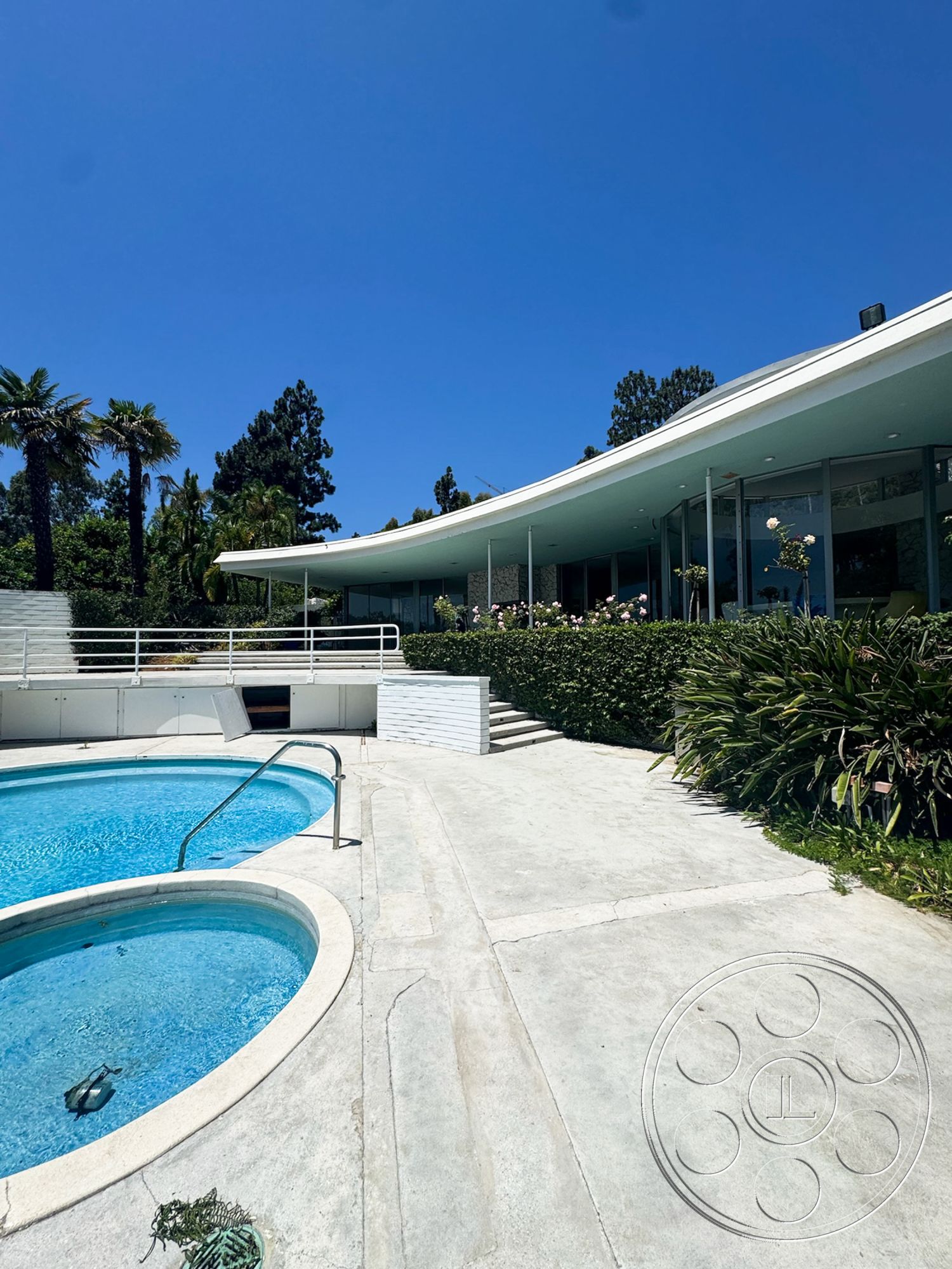 MCM 28 - exterior, landscaped garden, palm trees, glass windows, white railings, oval swimming pool, concrete patio flooring, minimalist architectural style, blue sky backdrop, steps leading to pool area, curved roof structure