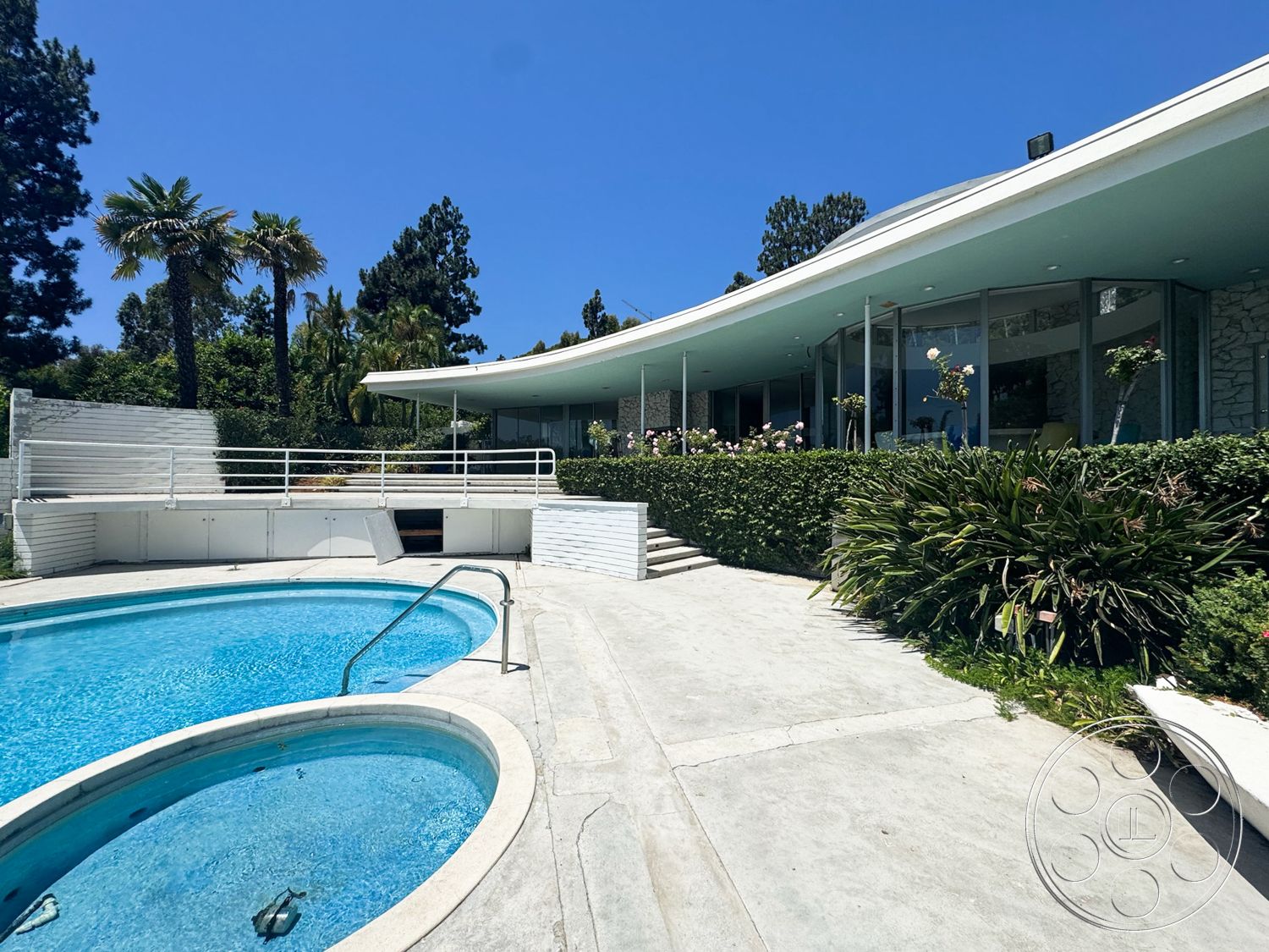 MCM 28 - exterior, stone accent wall, rectangular swimming pool, floor-to-ceiling windows, concrete steps, green hedges, landscaped garden area, concrete pool patio, palm trees in garden, blue sky view, curved house roof