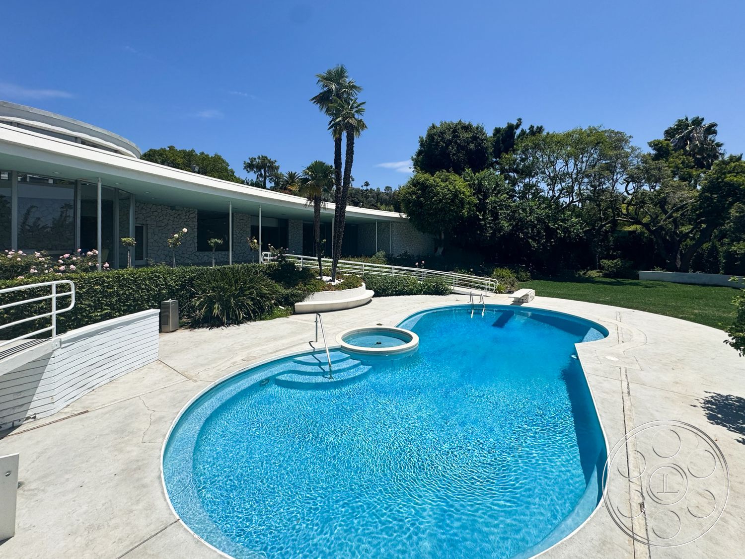 MCM 28 - exterior, palm trees, floor-to-ceiling windows, grass lawn, spa area, concrete pool deck, mid-century modern architecture, stone wall accents, oval swimming pool, landscaped garden area, horizontal roof overhang, white railing detail, curved geometric forms