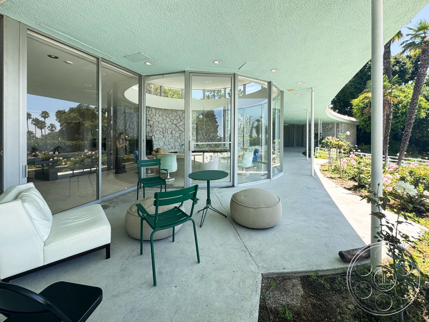 MCM 28 - exterior, glass sliding doors, steel framework, stone feature wall, open-air layout, palm tree landscaping, modern lounge seating, curved concrete patio floor, vaulted ceiling with textured finish, green garden patio, round ottoman seating, pedestal support columns, connecting indoor-outdoor spaces