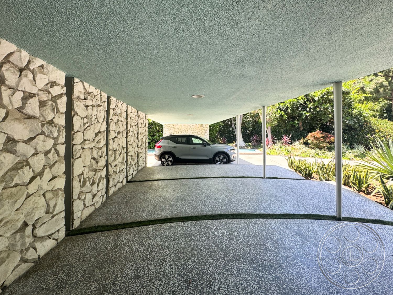 MCM 28 - exterior, smooth concrete surface, exposed stone wall, lush greenery setting, metal support columns, textured ceiling finish, curved driveway design, landscaped garden with succulents, pebble concrete driveway