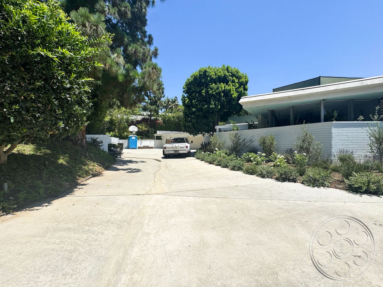 MCM 28 - concrete driveway, clear blue sky, tall pine trees, outdoor parking area, two-story home, white painted exterior walls, landscaped garden beds, modernist architectural style, bushy shrubs