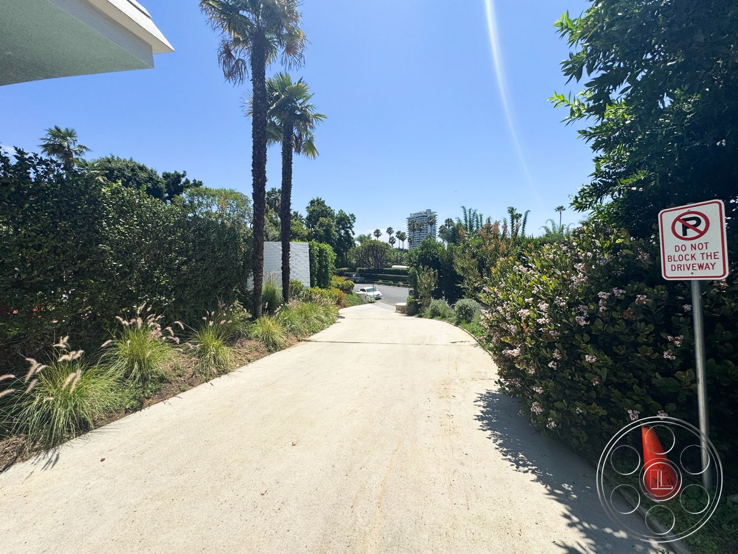 MCM 28 - exterior, lush greenery, palm trees, concrete driveway, curved driveway, clear blue sky, landscaped garden borders, decorative signage, pink flowering shrubs