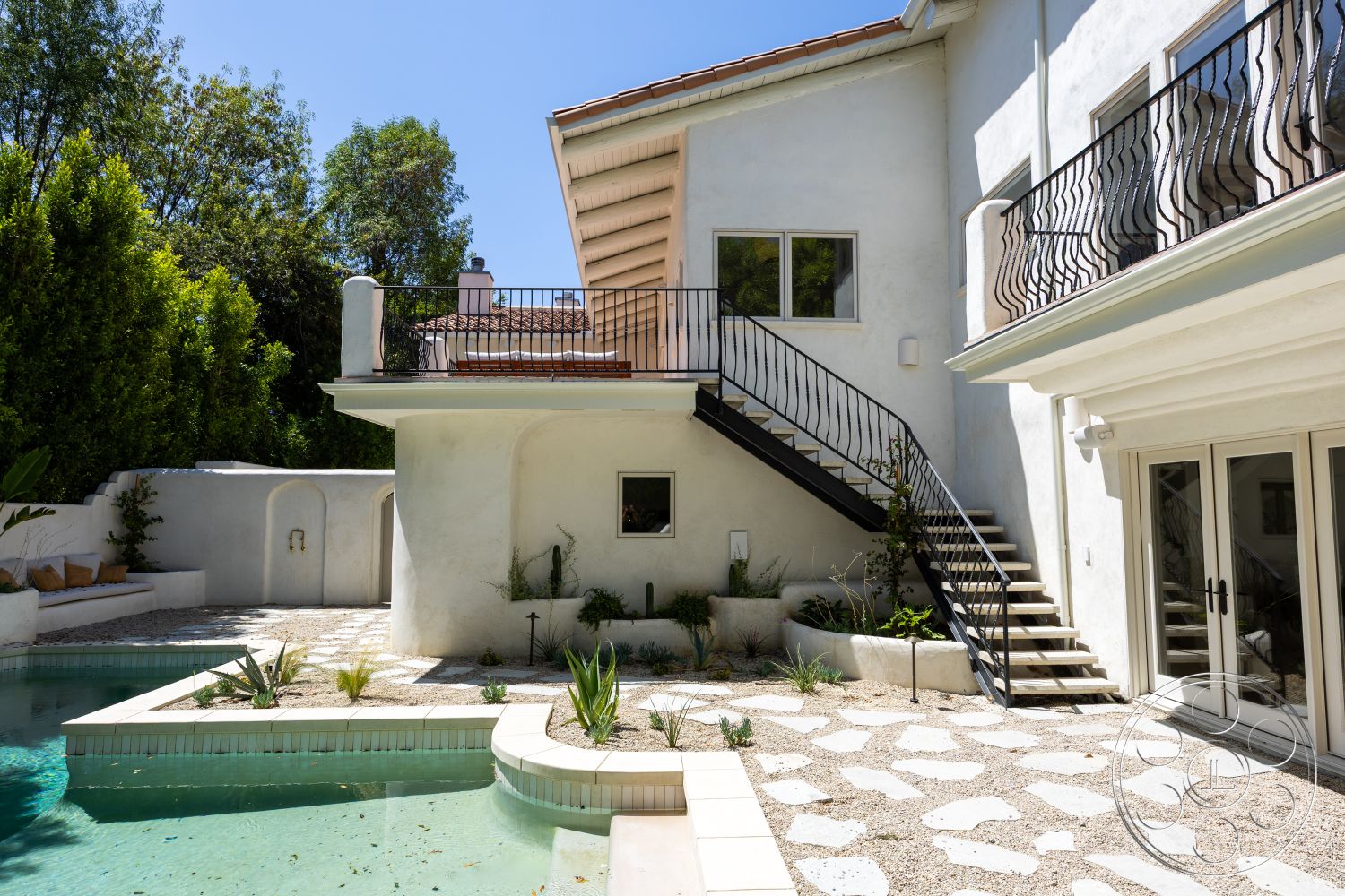 Medit 168 - exterior, outdoor seating area, rectangular swimming pool, modern Mediterranean architecture, concrete patio flooring, stucco wall finish, natural light through large windows, surrounding trees and greenery, irregular stone pavers, landscaped garden with succulents, upstairs balcony with wrought iron railings, wooden staircase with metal railings, sloped roof with tile shingles