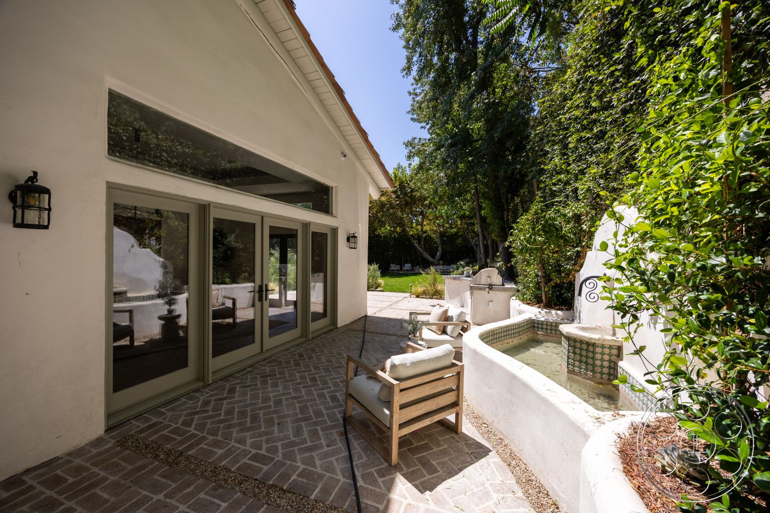 Medit 168 - exterior, white stucco walls, exposed wood ceiling beams, modern outdoor seating area, stone garden wall, lush greenery landscaping, herringbone brick patio flooring, green framed sliding glass doors, concrete fountain with tile accents, decorative lantern light fixture