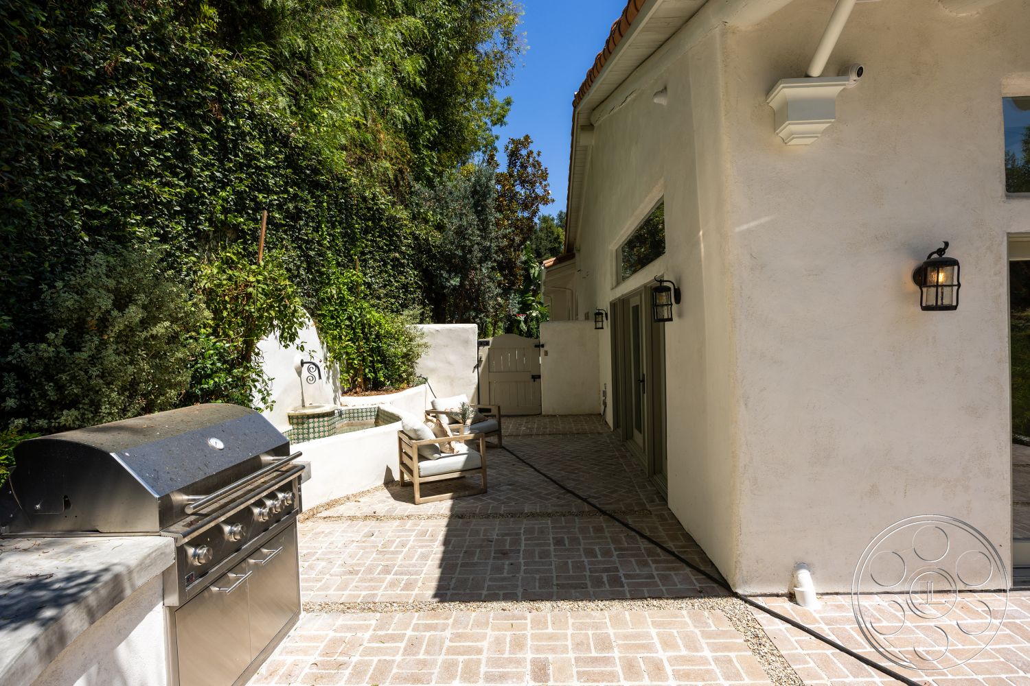 Medit 168 - exterior, transom windows, sloped terrain, decorative water feature, lush greenery backdrop, outdoor lighting fixtures, horizontal wooden fence, landscaped garden setting, stucco wall finish, brick pathway border, cobblestone patio flooring, stainless steel outdoor grill, built-in outdoor seating, architectural roof overhang